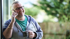 Stewart is leaning against a door frame. He is smiling, holding a mobile up to his ear and has a mug in his other hand. 