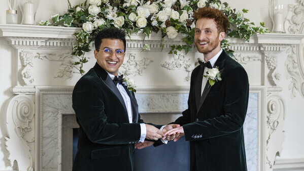 Two grooms in dark suits are standing in front of a white fire place. One groom is putting a ring on the other. There is a bouquet of white flowers on top of the fireplace.