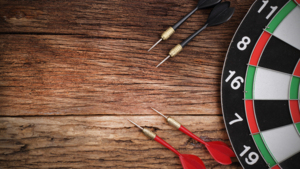 Four darts on a wooden table. Two are red and two are black.  Next to them is a darts board.