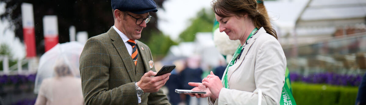 Two people are standing outside. The person on the left is wearing a chequered suit, blue and orange tie, blue hat and dark trousers. They are holding their phone in one hand and an umbrella in another. The person on the right is wearing a black and white patterned dress and white blazer. They are holding their mobile in both hands and have a green donation bucket hanging off their elbow.