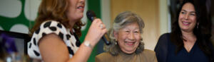 Three people are standing. A person with short brown hair wearing a black and white patterned dress is speaking into a microphone. To their right is a person with short hair who is wearing a brown cardigan and brown dress. They are smiling. Next to them is a person with dark hair and a blue dress. They are also smiling.