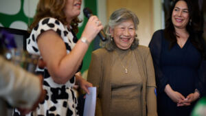 Three people are standing. A person with short brown hair wearing a black and white patterned dress is speaking into a microphone. To their right is a person with short hair who is wearing a brown cardigan and brown dress. They are smiling. Next to them is a person with dark hair and a blue dress. They are also smiling.