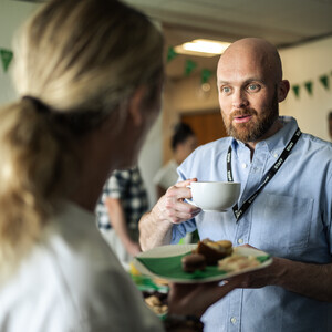 A person in a blue shirt and short, brown beard is holding a large white coffee cup in their hand. They are speaking to a person with long blonde hair wearing a white shirt. The person in the white shirt is holding a plate with food on it. They appear to be inside.