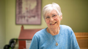 Ann has short white hair and is wearing a blue jumper. She is smiling and wearing a necklace with a gold chain and pendant. She appears to be inside.