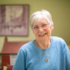 Ann has short white hair and is wearing a blue jumper. She is smiling and wearing a necklace with a gold chain and pendant. She appears to be inside.
