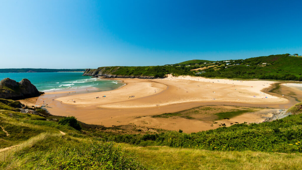 A view of a sandy beach from a grassy hillside. The sand is a light brown colour, the water is a bright blue, and the sky is clear and sunny.