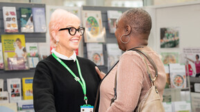 Two women talking, they are smiling the are standing in a healthcare setting with booklets behind them