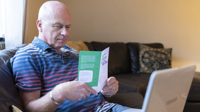 Steven sits on a sofa reading a cancer information leaflet. A laptop is open on his lap.