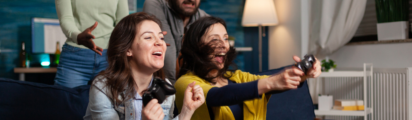 A group of people are in a house playing video games. One person in a yellow top is holding a gaming controller in their hands. Next to and behind them are people cheering. 