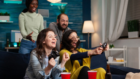 A group of people are in a house playing video games. One person in a yellow top is holding a gaming controller in their hands. Next to and behind them are people cheering. 
