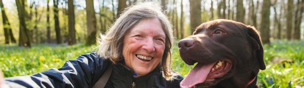 A woman with short hair is taking a selfie. She is with a brown lab. They are outside. The woman is wearing a blue coat. The lab has on an orange collar.