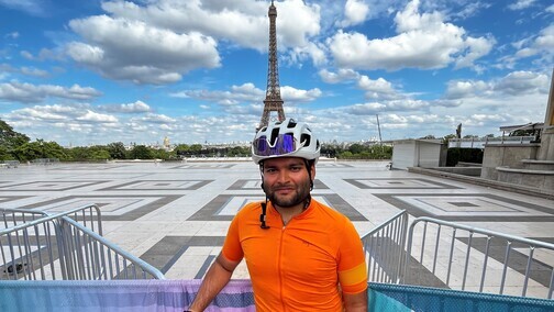 Nishad is standing in front of metal barriers outside. He is wearing a bicycle helmet, bright orange top, black trousers and blue trainers. In the distance behind him is the Eiffel Tower.