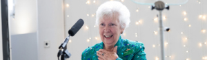 A speaker at a Thanks to You awards event. They have short white hair and are wearing a pink top and blue jacket. They are standing behind a stand that has a microphone on it. 