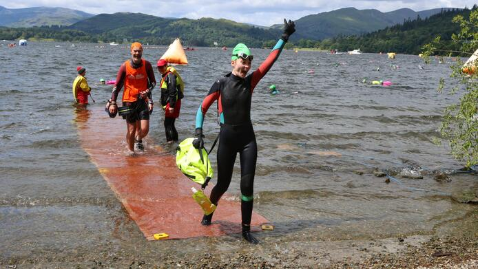 Heather is stepping out of lake onto a rocky beach. She has one arm in the arm and the other is holding a flotation device. Heather is wearing a wet suit, a swimming cap and googles.