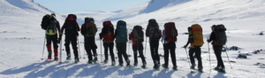 A group of hikes are walking in a row across a field of snow. They are wearing snow shoes and using walking sticks. They have hiking gear on and are wearing large rucksacks. They are outside in a large snow field.