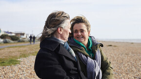 A couple sitting together on a bench by a pebble beach, with the sea in the background.