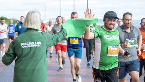 A Macmillan volunteer with a big green foam hand high fiving a Macmillan runner as they run past
