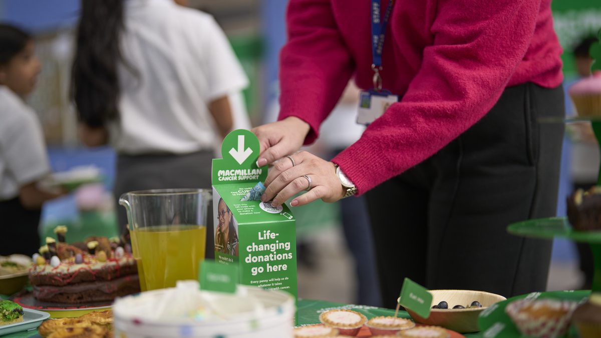 A person in a bright pink jumper is leaning over a table and placing a pound note in a Macmillan collection box. The box is on a table that has been decorated for a Coffee Morning. There are drinks and different sweat and savoury foods on the table. 