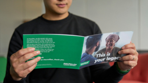 A person is holding open a Macmillan Gifts in Wills booklet. They are wearing a dark top. The booklets cover and the back of the booklet can be seen.
