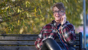 Karen is sitting on a park bench. They are speaking on their mobile phone. They are wearing a red, white and black patterned coat and black trousers. 