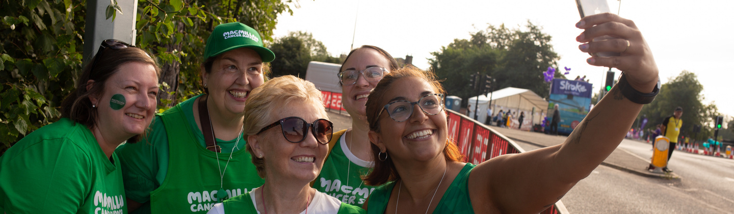 A group of Macmillan volunteers are taking a selfie. They are all wearing green Macmillan branded tops. They are standing outside.