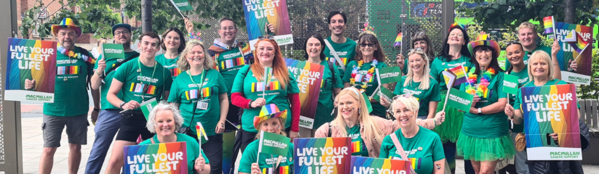 A large group of volunteers are at a Pride event. Some are holding up signs that say live your fullest life. They are wearing green Macmillan branded tops.