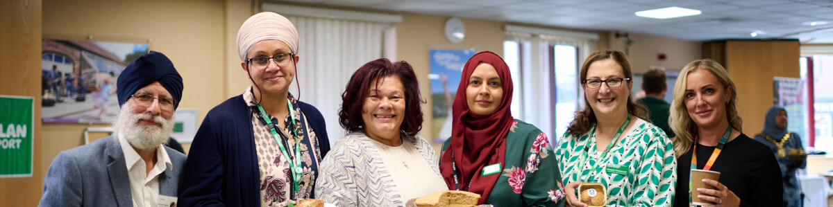 Six people standing behind a Macmillan Coffee Morning table filled with cakes and pastries. They are each holding a cake or drink.