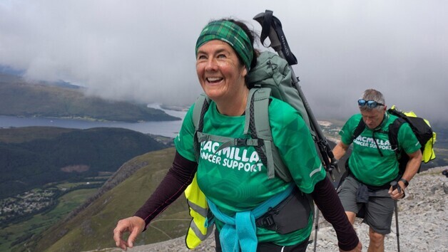 Two people are hiking along a rocky path. They are both wearing green Macmillan branded tops. They are also wearing other hiking gear like backpacks. 