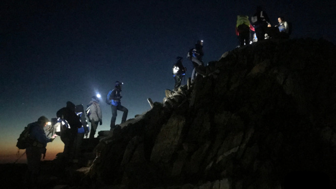 Hikers walking at night up a rocky terrain. They are wearing different hiking gear, including head lamps.