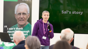 Sal is standing on a stage and is speaking into a microphone. She is wearing a dark purple hoodie and blue jeans. Behind Sal is a large screen with the words Sal's story on it. There is also a Macmillan branded banner. There is a crowd of people sitting and listening to Sal's story.