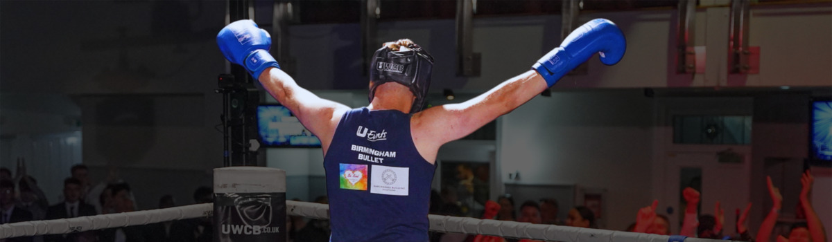 A person wearing boxing gloves has their arms in the air and is facing away from the camera. They are in a boxing ring and people are cheering for them. They are also wearing a blue top, dark shorts, and a helmet to protect their head when boxing.