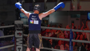 A person wearing boxing gloves has their arms in the air and is facing away from the camera. They are in a boxing ring and people are cheering for them. They are also wearing a blue top, dark shorts, and a helmet to protect their head when boxing.