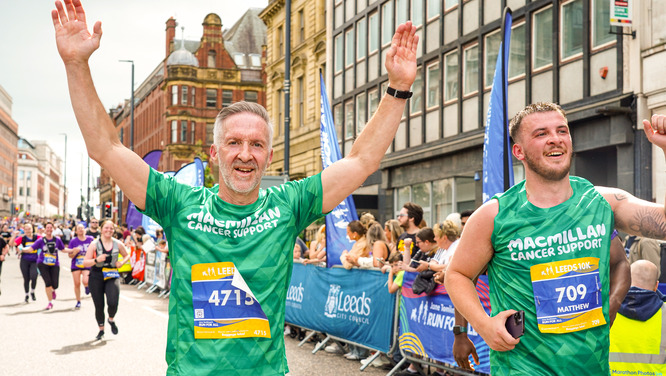 Two men are running in the Leeds 10K. Both are wearing green Macmillan branded running tops and black trousers. The man on the left has his arms in the air. The one on the right is waving to onlookers.