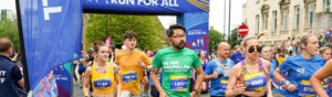Runners taking part in the Leeds 10K event. One runner is wearing a green Macmillan branded top and black shorts. There are onlookers on either side of the runners behind barriers.