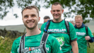 Three men are walking in a row. The man in front is smiling. All three men are wearing green Macmillan branded tops and ruck sacks. They appear to be hiking outdoors.