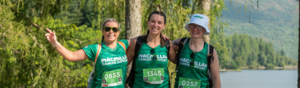 Three hikers are standing side to side with their arms around each other. They are hiking on a gravel path near a lake. They are all wearing green Macmillan branded tops, a event ID card attached to the top and wearing black leggings.