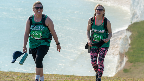 Two hikers are walking up a grassy slope near a beach. They are both wearing green Macmillan tops and other hiking gear. They have event ID badges attached to their tops. 
