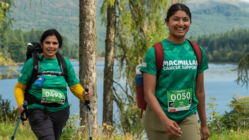 Two hikers are walking along a path near a lake. They are both wearing green Macmillan branded tops with event ID badges pinned to the tops. The hiker on the left is using walking sticks. 