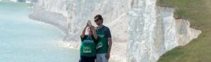 Two hikers are posing and taking a selfie. They are standing on a grassy slope near a beach with white cliffs. Both hikers are wearing green Macmillan branded tops and have event ID badges pinned to their tops. 