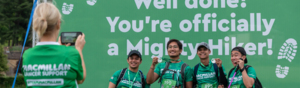 A Mighty Hike volunteer is taking a photo on mobile of a group of hikers standing in front of a large banner. Two of the hikers are holding up finisher's medals. The banner behind then states: 'Well done! You're officially a Mighty Hikers!' 