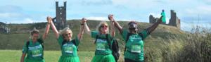 A group of four hikers are walking holding hands along a grassy path. Three of the hikers are wearing green tutus. Behind them are grassy fields and old ruins.