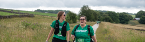 Two hikers are walking next to each other on a gravel path. The are both wearing green tops, dark shorts and sunglasses. The path goes through a grassy field. 