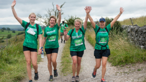 Four hikers are walking side by side along a gravel, rocky path. They have their arms in the air. All of them are wearing a green top and dark shorts. The path goes along a grassy field.
