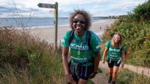 Two people are hiking on a sandy path near the beach. Both hikers are wearing green tops, dark shorts and rucksacks.