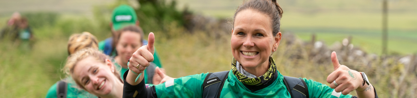 Women walking through the countryside wearing green Macmillan tops. The woman at the front is smiling and putting both thumbs up.