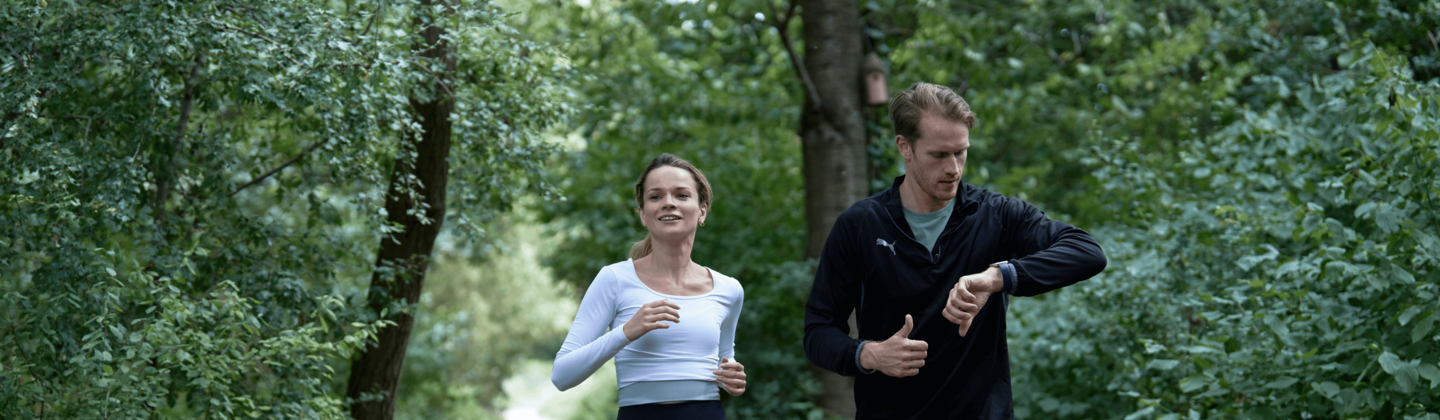 Two people are running along a dirt path in the woods. The person on the left is wearing a white top and dark trousers. The person on the right is looking at their watch. They are wearing a dark top and dark shorts.