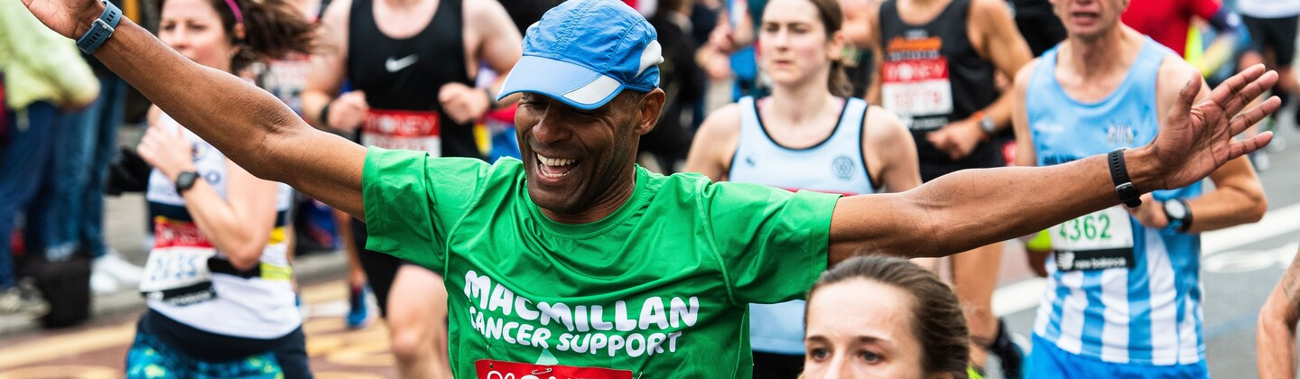 Man wearing a green Macmillan top and a blue hat with his arms spread out wide and smiling while running at the London Marathon