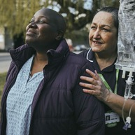 A Macmillan nurse standing behind a Black patient outside with her hands on her shoulders.