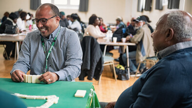 Two men are playing dominoes on a table inside a large conference room. Behind them are other tables with people playing dominoes and chatting. They are all members of the Pineapple Club in South East London.