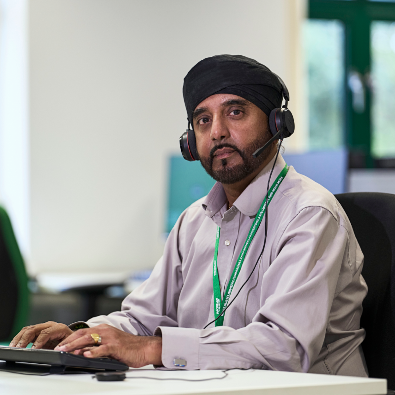 A photo of Akbal, a person who works on the Macmillan Support Line. Akbal is seated at a desk and typing into a computer keyboard, while wearing a phone headset.